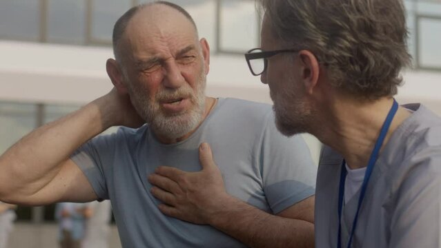 Health care specialist sits with aged patient on the bench and discusses his treatment. Elderly man talks with doctor and complains of neck pain. Mature professional in medical uniform works outdoor.