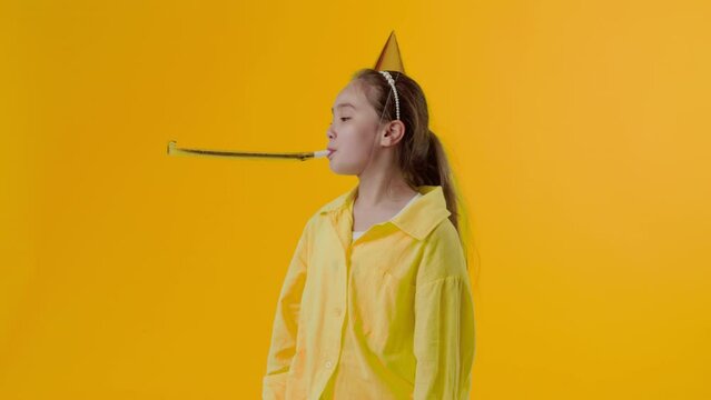 Young asian girl in the paper cap blowing a party horn and looking at the camera with an unhappy face on a yellow background. Concept of lack of interesting activities for a child at the party
