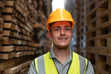 Male warehouse worker wearing uniform and helmet safety standing and smiling in plank wooden warehouse. The factory makes pallet wood and export.
