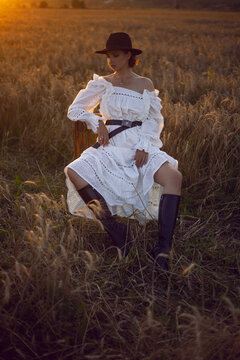 fashionable village woman in a white dress, boots and a black hat sits on a chair in a wheat field at sunset in summer.