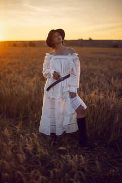 fashionable village woman in a white dress, boots and a black hat stand in a wheat field at sunset in summer.