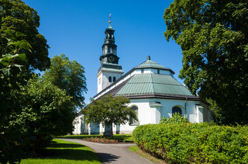 Köping church, white paint facade, copper-covered gable roof, Baroque style spire on copper-clad tower with clock, Old religious building surrounded by green garden, Sweden