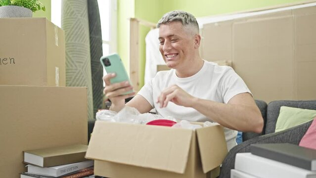 Young caucasian man unpacking cardboard box using smartphone at new home