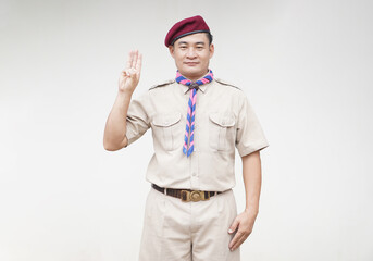 Handsome Asian man wear boy scout uniform, red cap, blue and pink striped scarf, make hand sign symbol of scout. Concept, educational career with uniform in school, Thailand.   - obrazy, fototapety, plakaty