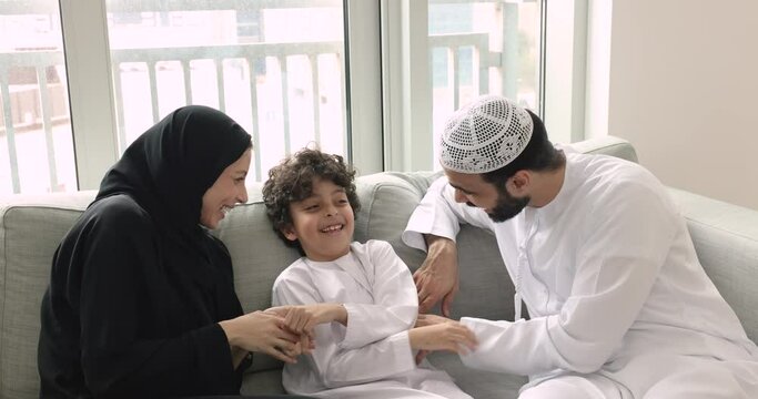 Loving mother in black hijab spend time with family tickling little 5s son sit on sofa with husband in white traditional thobe enjoy playtime on carefree weekend leisure at home. Family bond, culture