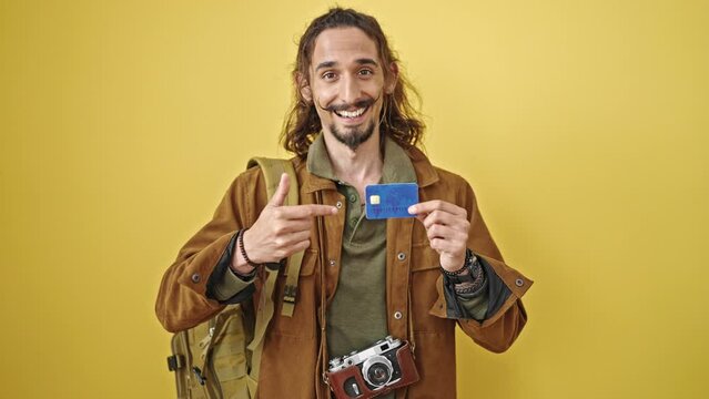 Young hispanic man tourist pointing to credit card doing thumb up gesture over isolated yellow background