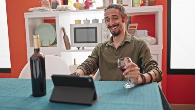 Young hispanic man drinking glass of wine watching video on touchpad at dinning room