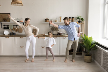 Happy active mom, dad and cute toddler kid dancing in spacious home kitchen, celebrating moving...