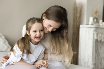Joyful young mother playing with toddler child on bed, cuddling, tickling cute laughing kid, holding sweet little daughter in arms, enjoying motherhood, family leisure, funny activities