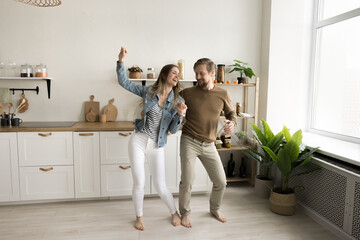 Positive active couple dancing together in spacious new home kitchen, laughing, celebrating anniversary, real estate buying, apartment rent after moving, enjoying music, party for two
