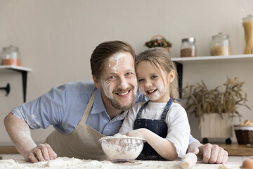 Cheerful funny little daughter and dad posing in home kitchen with floury faces, looking at camera,...