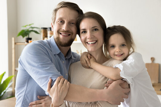 Happy cute toddler kid and dad embracing cheerful mom with love, gratitude, affection. Positive parents and little daughter girl hugging at home, looking at camera with toothy smiles. Family portrait