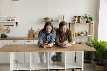 Positive digital addicted couple standing close in kitchen, using gadgets separately, avoiding...