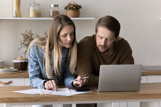 Focused married couple counting mortgage fees on calculator, using laptop at kitchen table, checking financial legal documents, real estate agreement. Husband and wife doing accounting work at home