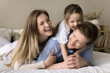 Obraz na płótnie Canvas Joyful excited family having fun at home. Happy young parents and little toddler kid stacking on bed, playing in bedroom, enjoying leisure, weekend, activity, parenthood, childhood