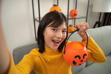 Young asian woman holding smartphone and talking with pumpkin toy in hand at living room. She preparing to join in halloween party.