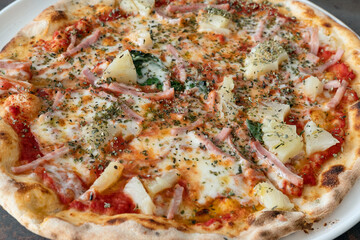 Close-up of a whole pineapple and ham thin crust pizza on a plate abundant in delicious ingredients and sprinkled over with flavorful dried oregano, traditional Italian dish baked in a wood-fired oven