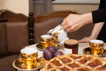 A girl pours milk to coffee in retro cups gilded on old wooden table next to a home-baked cake with plum jam, retro atmosphere. Coffee is even tastier in such cups
