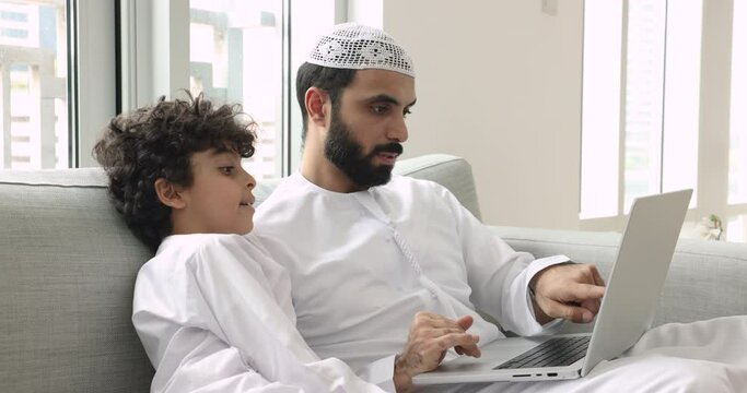Muslim man teach little son to use laptop, family discuss purchase via web store sit on sofa with wireless computer talk about new app for hobby. E-shopping, educational website for kid development