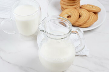 Milk in a transparent glass and jar on a marble table. Round ring shaped German spritz biscuits at background