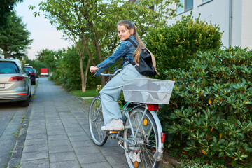 Schoolgirl child 10, 11 years old with backpack on bicycle on street near house