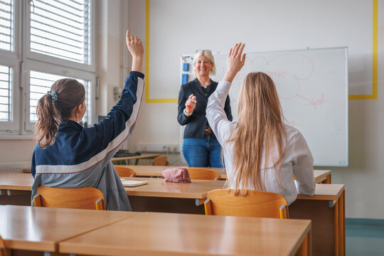 Girl student writes down notes and raises her hand while a female teacher lecturing in the class in front of the blackboard. Concept of classroom environment.