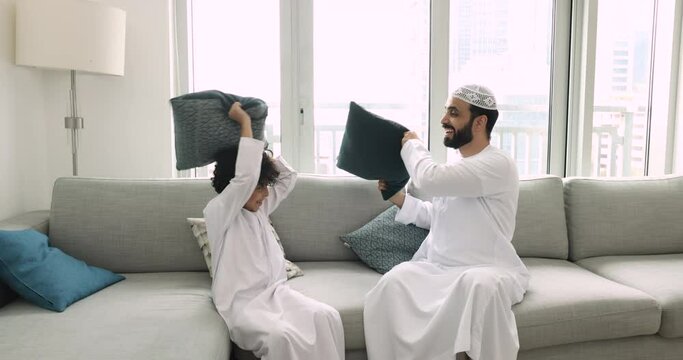 Happy Muslim father and cheerful preschooler son in traditional wear playing pillow battle seated on couch in living room, loving family having fun, enjoy playtime and communication on weekend at home