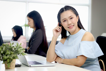 Portrait of happy beautiful Asian woman officer sitting and working at office desk with blurred background of her busy working colleagues. Pretty attractive smiling female staff worker at workplace.