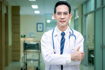 Portrait of confident smiling senior Asian male doctor in white workwear holding stethoscope and looking at camera, standing with arms crossed in corridor of medical clinic, happy doctor at hospital.