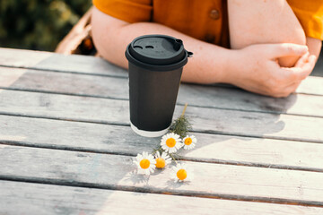 Black paper cup for coffee or tea standing on a wooden table with a sprig of daisies and woman's hands in the background