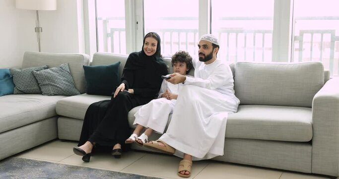 Muslim family with son watching television at home. Young woman in hijab spend weekend leisure with husband and son in traditional islamic wear laughing enjoy cool comedy movie feel happy. Pastime, TV
