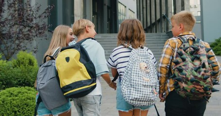 Group of four smart diverse classmates with colorful backpacks are having fun talking while going to school school, outdoors. Back view.