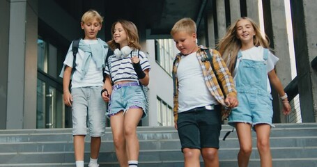 Front view of group of four attractive caucasian schoolchildren walking together down the stairs in college campus talking and laughing with joy. Education and school children life concept
