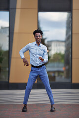 Happy African American man dancing on city street near building, smiling and wearing formal clothes blue trousers and shirt. Handsome male model outdoors