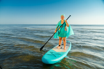 A woman in a turquoise swimsuit with a skirt and a scarf on her head on a SUP board swims to the seashore.