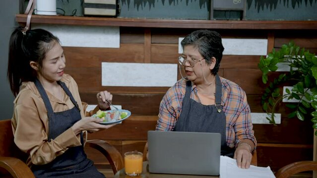 Retired woman elderly mother sits in cafe for daughter. Wear glasses to see the computer screen better. daughter walks holding a plate of fruit glass of orange juice for mom for freshness during work