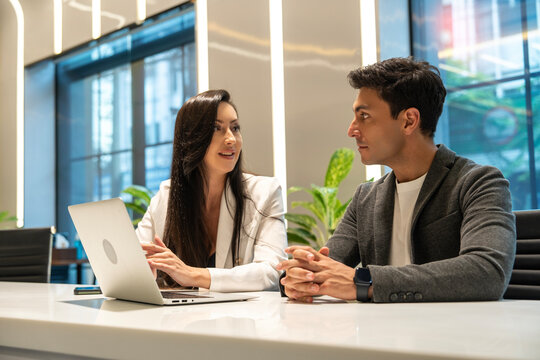 Caucasian young woman secretary wearing a suit sits typing on a laptop and discusses data problems in the system with a male executive about working in a conference room