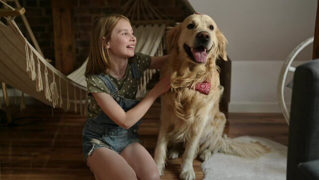 Girl child with golden retriever dog sitting at floor together at home with hammock. Pretty preteen kid hugging purebred doggy pet in loft room