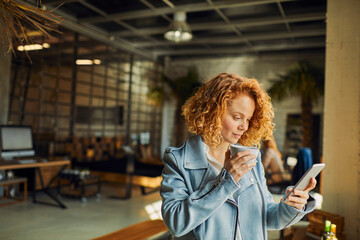 Young Caucasian woman drinking coffee and using a smartphone in a startup company office