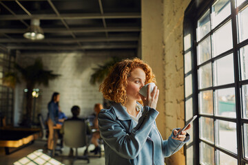 Fototapeta premium Young Caucasian woman drinking coffee and using a smartphone in a startup company office