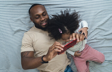 Happy african american father lying on cozy bed showing mobile application, video game or online cartoons to funny cute little baby daughter. Black family having fun enjoy funny moments in bedroom.