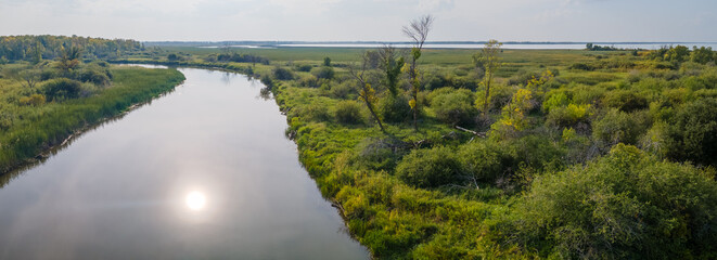 Panoramic view of a large calm river that is surrounded by grasses and shrubs that have yellowish green colors resulting from autumn weather.  The sun is reflected in the water.  
