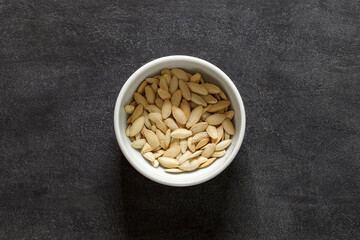 Pumpkin seeds, unpeeled with salt in white bowl, on dark background, top view, space to copy text