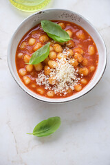 Bowl of tomato gnocchi soup served with fresh basil and grated parmesan, vertical shot on a light-beige marble background, elevated view