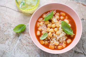 Tomato soup with potato gnocchi, grated parmesan and green basil on a pinkish granite background, elevated view, horizontal shot