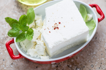 Feta cheese with fresh basil leaves in a red serving pan, middle close-up on a beige stone surface, horizontal shot