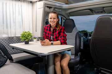 Young adult brunette woman wearing plaid shirt sitting inside the motor home while parked in a camper van parking place. RV rental.