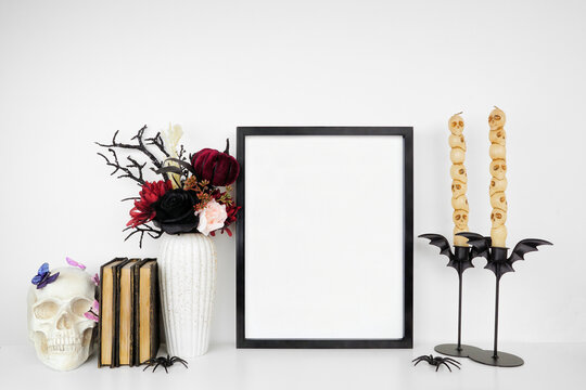 Halloween gothic romance mock up. Black frame on a white shelf with red and black flowers, skull, books and candles. Portrait frame against a white wall. Copy space.