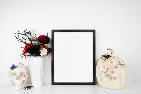 Halloween gothic romance mock up. Black frame on a white shelf with red and black flowers, skull and floral pumpkin. Portrait frame against a white wall. Copy space.