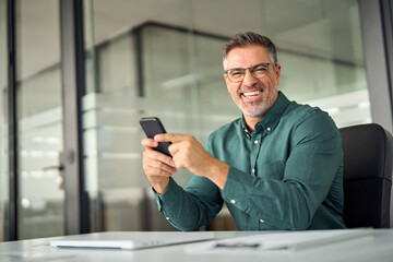 Obraz na płótnie Canvas Cheerful older professional businessman, happy middle aged business man entrepreneur laughing looking at camera holding smartphone using cell phone mobile technology sitting at work desk in office.